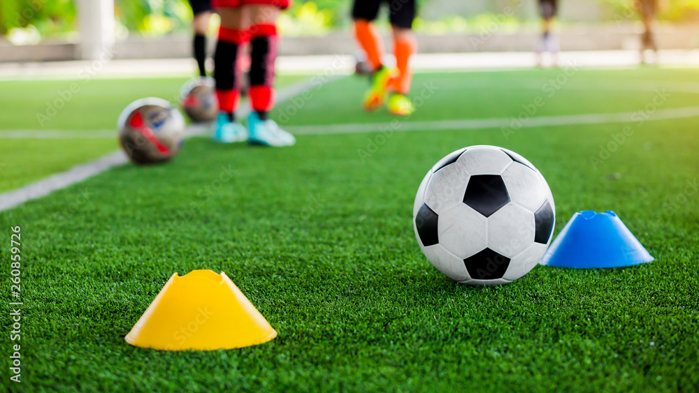 football between marker cones on green artificial turf with blurry soccer team training, blurry kid soccer player jogging between marker cones and control ball with soccer equipment in soccer academy.