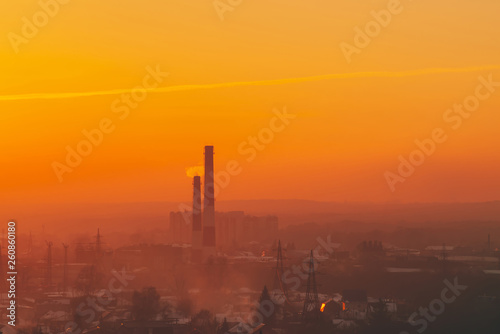 Smog among silhouettes of buildings on sunrise. Smokestack in dawn sky. Environmental pollution on sunset. Harmful fumes from stack above city. Mist urban background with warm orange yellow sky. © Daniil