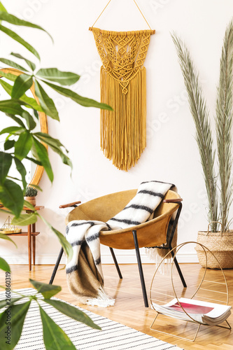 Stylish and minimalistic scandi interior of living room with design gold armchair, dressing table with mirror, plants, palm leaves, yellow macrame and accessories. Warm and cozy space of home decor. 