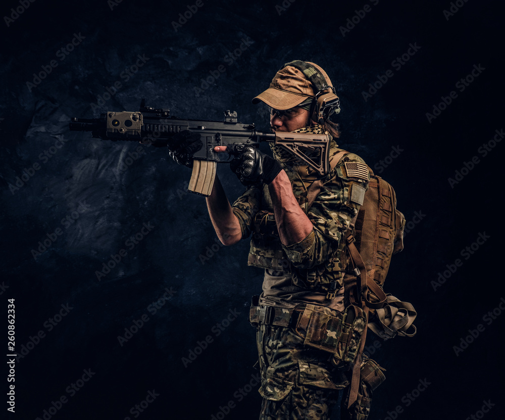 The elite unit, special forces soldier in camouflage uniform holding an assault rifle with a laser sight and aims at the target. Studio photo against a dark wall