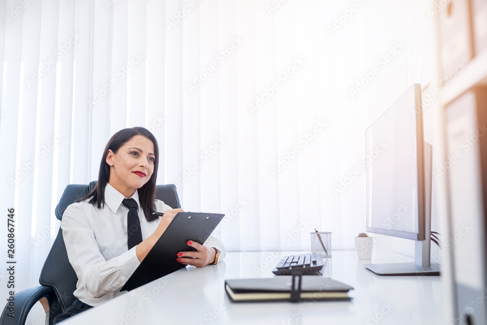 Smilling young business woman sitting in her office and writting on her clipping board. Smilling young business woman sitting in her office and writting on her clipping board.