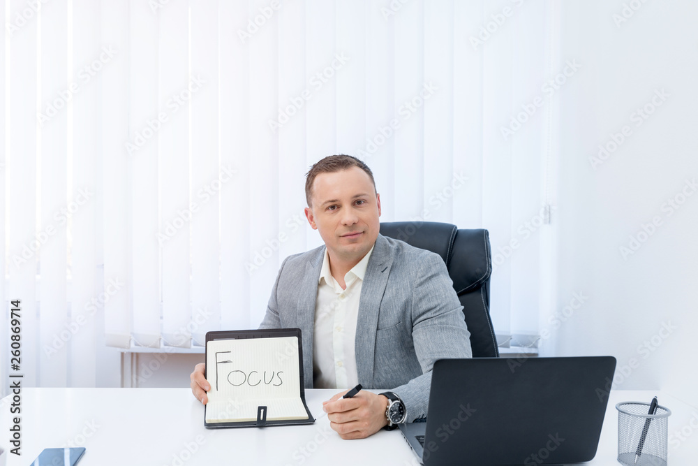 Young successful businessman showing Focus! written in the notebook while sitting behind an office desk in the office. Young successful businessman showing Focus! written in the notebook while sitting behind an office desk in the office.