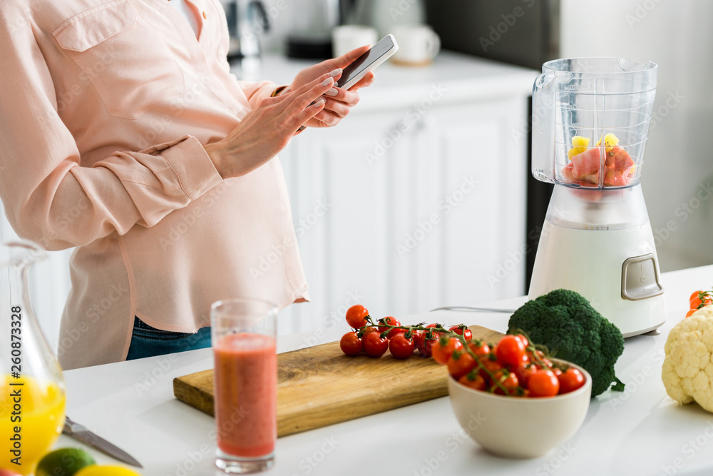cropped view of pregnant woman using smartphone in kitchen near cherry tomatoes on cutting board cropped view of pregnant woman using smartphone in kitchen near cherry tomatoes on cutting board
