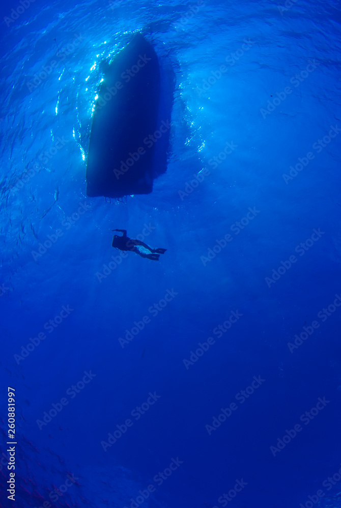Fototapeta premium A silhouette shot of divers returning to a dive boat. The water is very clear and the sun can be seen behind the dive boat.