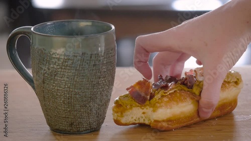 Woman eating a maple bacon donut for breakfast with coffee. The lady enjoys the sweet and savory pastry with her morning latte. The specialty gourmet doughnut flavor is to celebrate National Donut Day