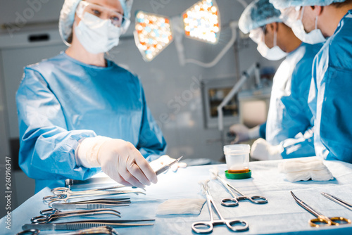 selective focus of doctors doing operation and nurse in uniform and medical cap holding scalpel photo