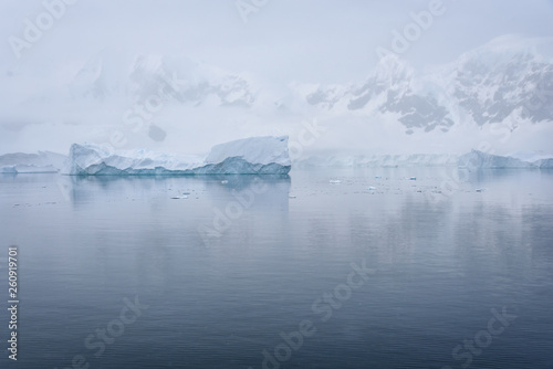 Tabular iceberg floating in a serene Paradise Harbor, against a foggy mountain background, Antarctica