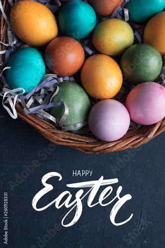 Happy Easter hand lettering inscription and colorful easter eggs in basket on black wooden background.