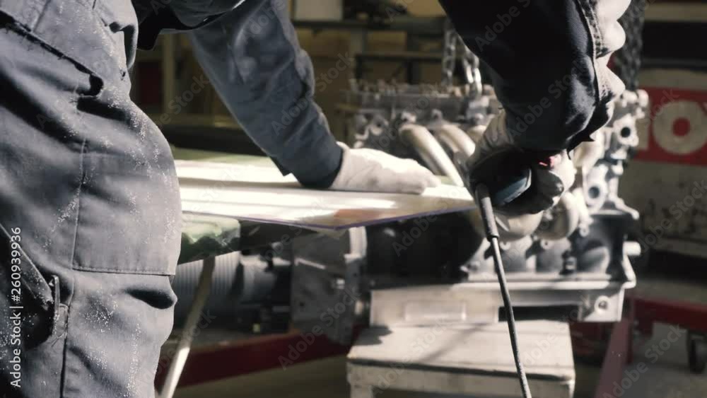 Car mechanic using circular polishing drill power tool to polish metal plate in sunny dusty service. Car engine in background. Car mechanic using circular polishing drill power tool to polish metal plate in sunny dusty service. Car engine in background.