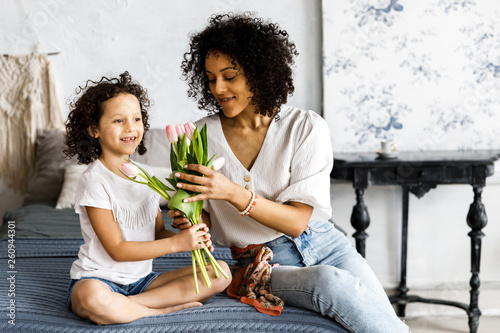 Mom and little cute curly girl sitting on bed with tulips in their hands