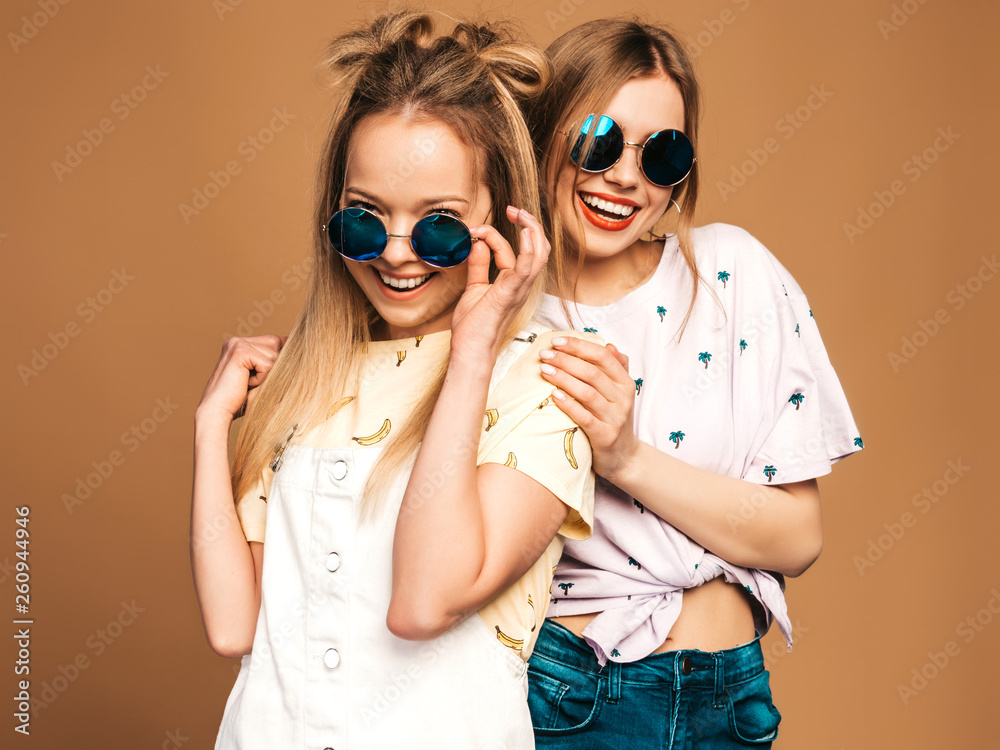 Two young beautiful smiling blond hipster girls in trendy summer colorful T-shirt clothes. Sexy carefree women posing near beige wall in round sunglasses. Positive models having fun