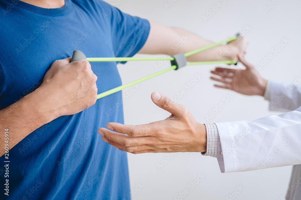 Doctor physiotherapist assisting a male patient while giving exercising treatment on stretching his arm with exercise band in the clinic, Rehabilitation physiotherapy concept