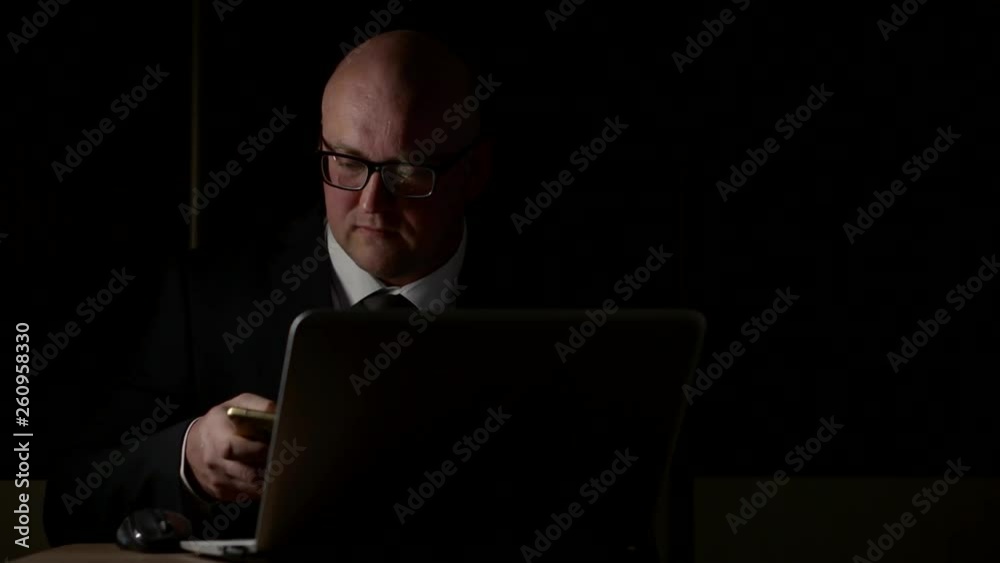 The business owner is a man in glasses and a suit working on his personal computer and mobile phone. He works late in an empty office. The business owner is a man in glasses and a suit working on his personal computer and mobile phone. He works late in an empty office.