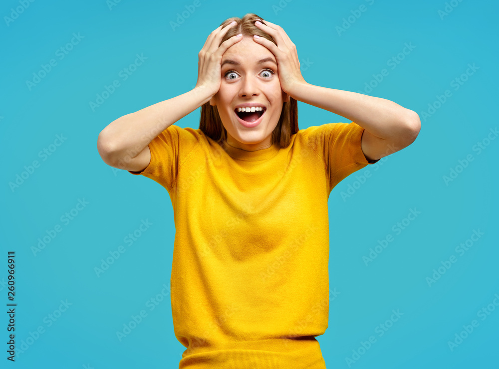 Overjoyed girl holds hands on head and looks joyfully at camera. Photo of girl in yellow sweater on blue background. Emotions and Omg concept