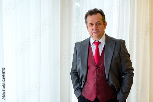 Portrait of successful senior business man 60 years old, dressed in suit and red vest holds hands in pockets on windows background. Confident and intelligence