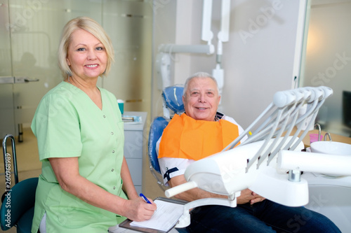 Dentist woman and senior man patient looking together to camera in dental clinic. Dental care for elder people. Dentistry, medicine and health care concept
