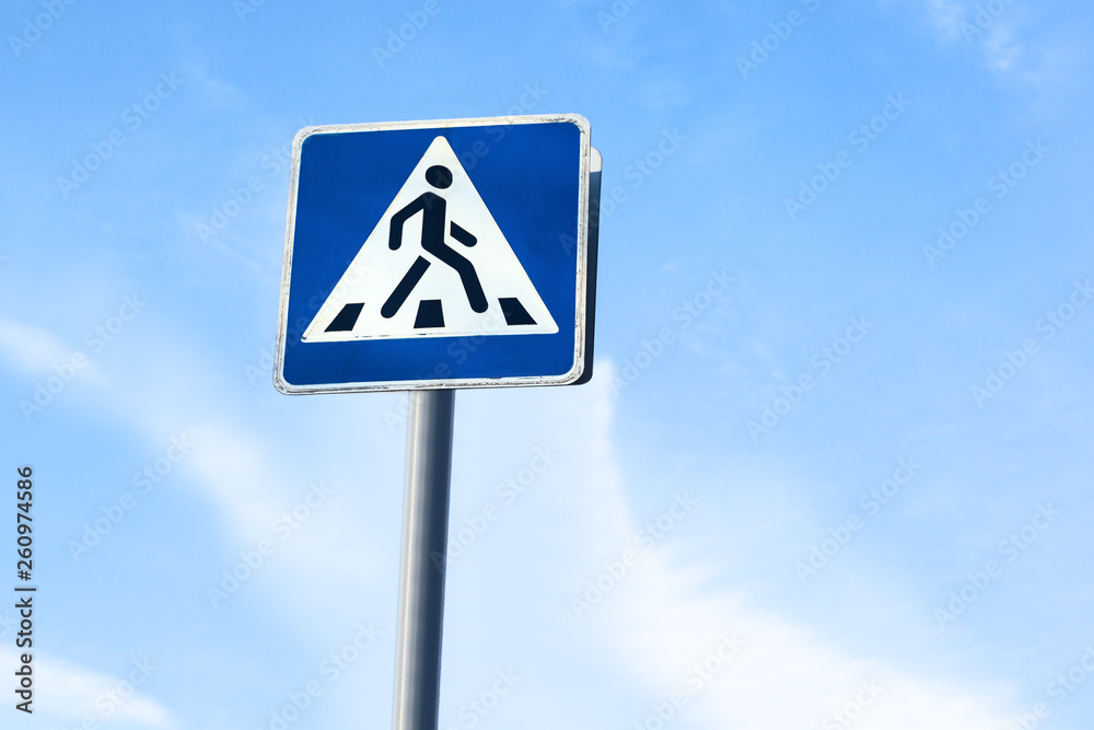 Road sign - Pedestrian crossing. Square blue and white metal plate with white stripes and a walking man. Blue sky background. Road sign - Pedestrian crossing. Square blue and white metal plate with white stripes and a walking man. Blue sky background.