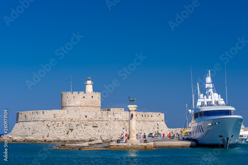 Rhodes, Greece - August 2016: Hirschkuh statue at the entrance in the Mandraki old harbour, with Fort of Saint Nicholas in background. Stone historical building on the seashore against clear blue sky. © Creatikon Studio