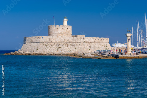 Rhodes, Greece - August 2016: Hirschkuh statue at the entrance in the Mandraki old harbour, with Fort of Saint Nicholas in background. Stone historical building on the seashore against clear blue sky.