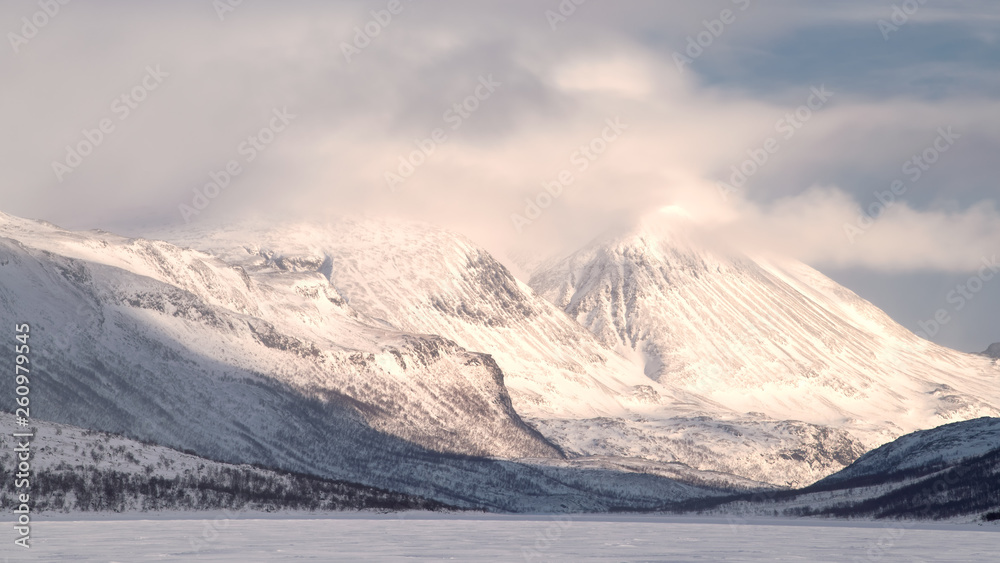 Swedish Lapland mountain landscape in winter with snow and cloud covered peak Kuoperatjakka in Sjaunja Naturreservat during sunset with frozen lake in foreground Swedish Lapland mountain landscape in winter with snow and cloud covered peak Kuoperatjakka in Sjaunja Naturreservat during sunset with frozen lake in foreground