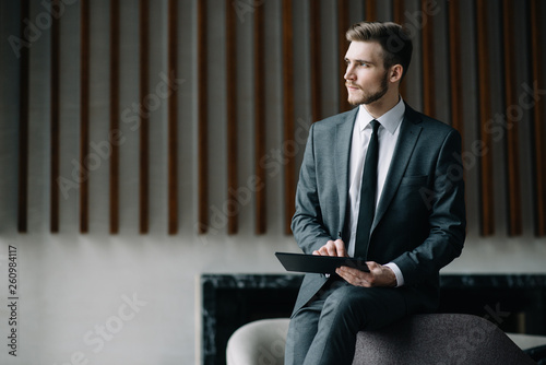Young handsome man sitting in office with cup of coffee and working on project connected with modern cyber technologies. Businessman with notebook trying to keep deadline in digital marketing sphere.