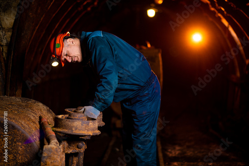 An elderly man dressed in work overalls and a helmet stands near the old inverted vogonetki. Mine worker. Miner