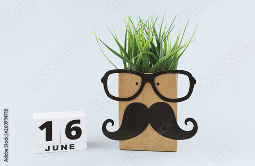 Greeting card with cheerful funny face on paper bag with glasses and mustache, and hair from grass and save date white wooden block calendar for 16st June to celebrate Father's day