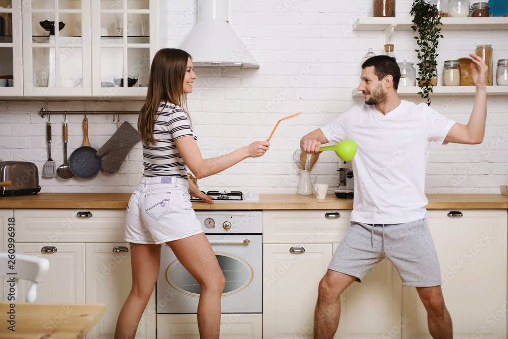 Funny couple fight with utensils tools while cooking at home together, husband and wife having fun feeling playful holding kitchenware struggling in the kitchen preparing healthy food Funny couple fight with utensils tools while cooking at home together, husband and wife having fun feeling playful holding kitchenware struggling in the kitchen preparing healthy food