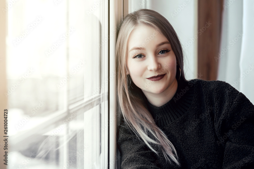 beautiful young girl sitting alone on the windowsill and looking at the camera and smiling. beautiful young girl sitting alone on the windowsill and looking at the camera and smiling.