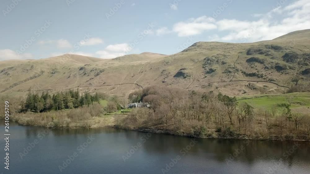 Aerial panning shot across Lake district national park, mountain range in background. White rural home in between dense woodlands alongside lake. Aerial panning shot across Lake district national park, mountain range in background. White rural home in between dense woodlands alongside lake.