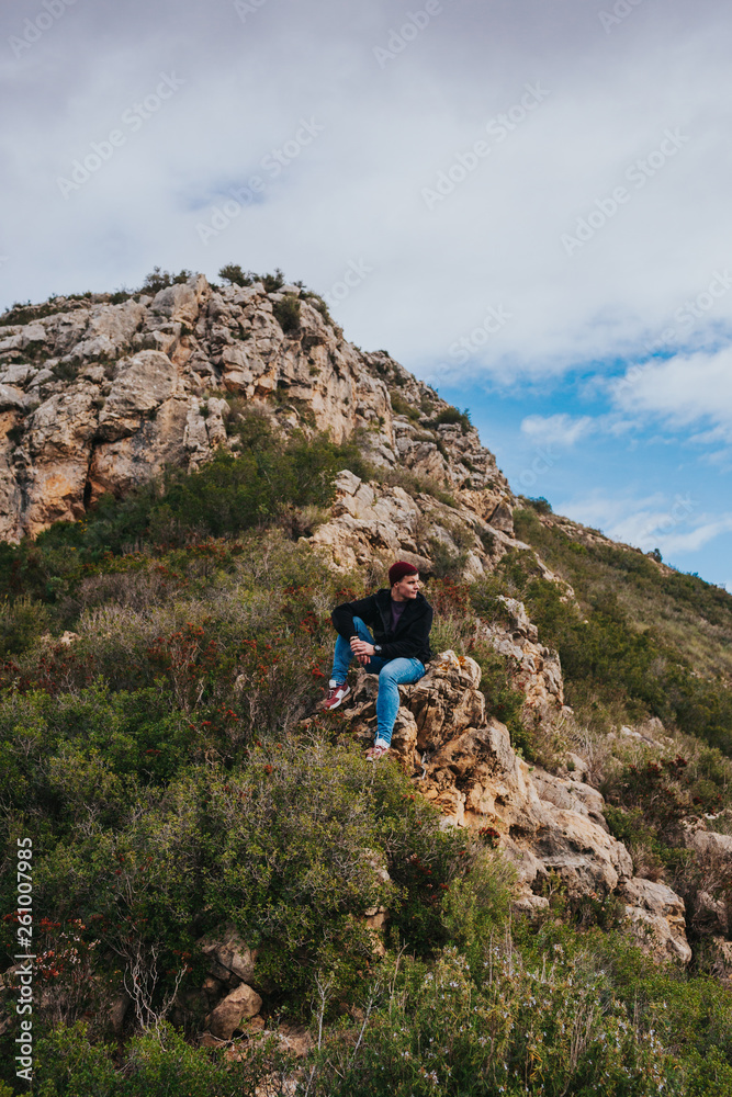 Young male sits on the rock and enjoys beautiful mountain landscape.