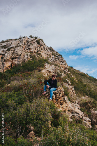 Young male sits on the rock and enjoys beautiful mountain landscape.