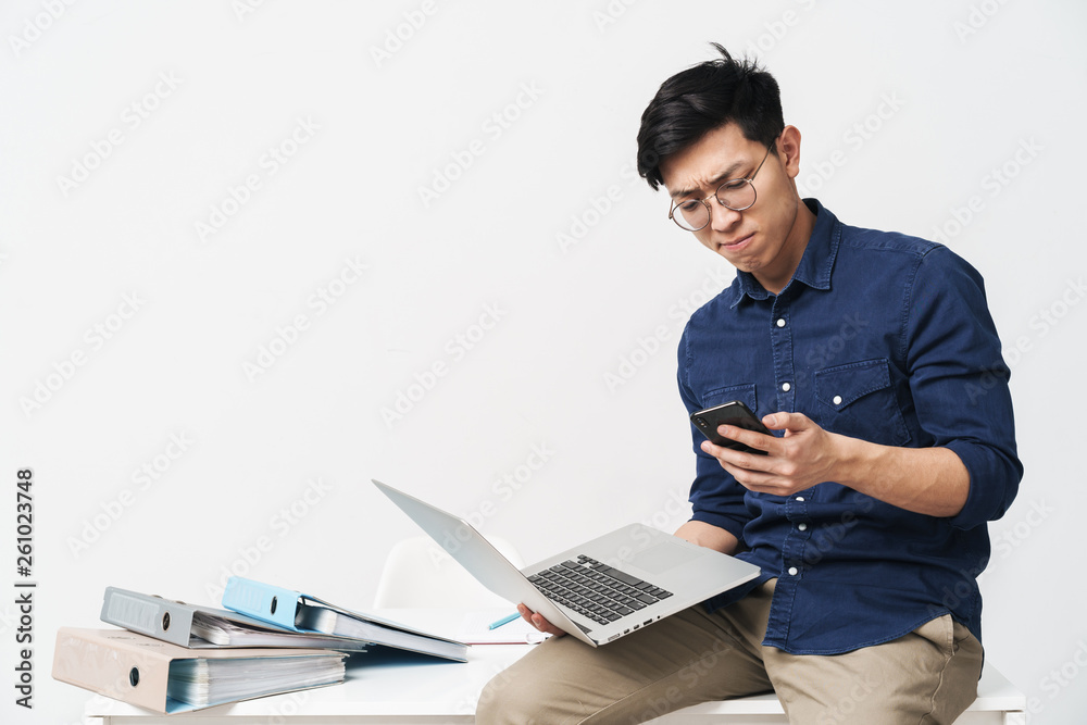 Φωτογραφία Photo of serious asian man 20s wearing eyeglasses holding smartphone and laptop