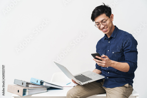 Photo of smiling asian man 20s wearing eyeglasses holding smartphone and laptop while working in office