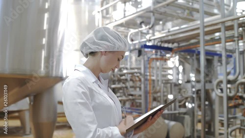 Female Food factory worker monitors production and takes notes on tablet at modern diary factory. Milk processing line