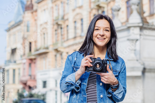Portrait of a positive smiling caucasian hipster woman in blue jeans jacket taking picture with an old-fashion camera looking at camera outdoor with city street on background.
