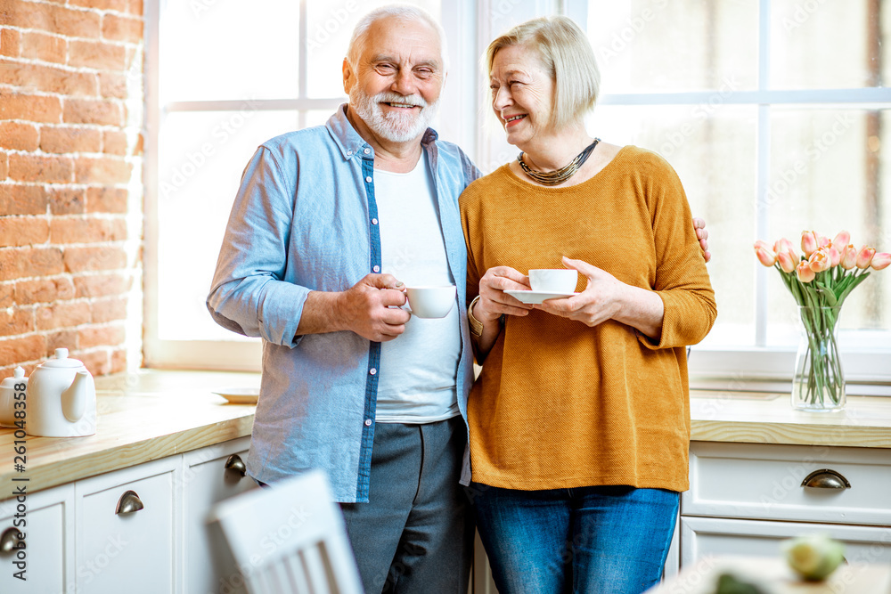 Fototapeta premium Portrait of a lovely senior couple talking together, standing with coffee cups near the window at home