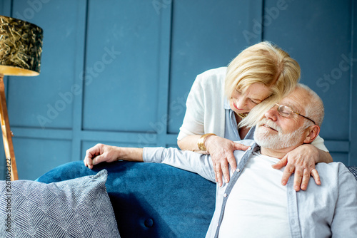 Portrait of a lovely senior couple dressed casually embracing together on the couch at home on the blue wall background