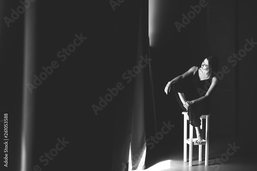 The social theme female loneliness pain suffering. Abastration male violence family. A young beautiful Caucasian woman in black clothes sitting on three chairs in large dark room. Srach and despair