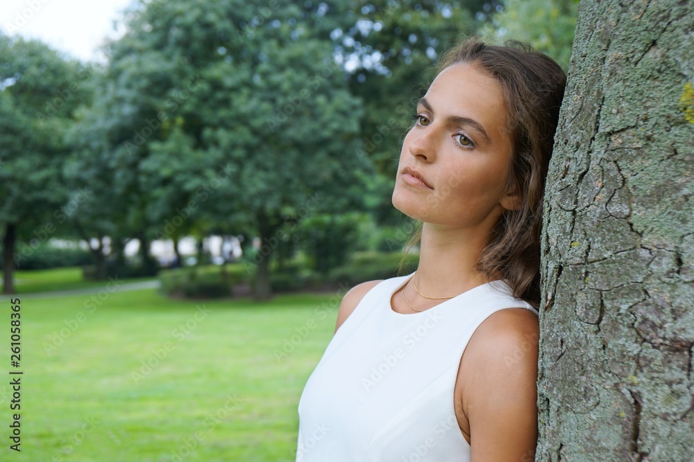 sad young woman leaning against tree gazing absentmindedly                              