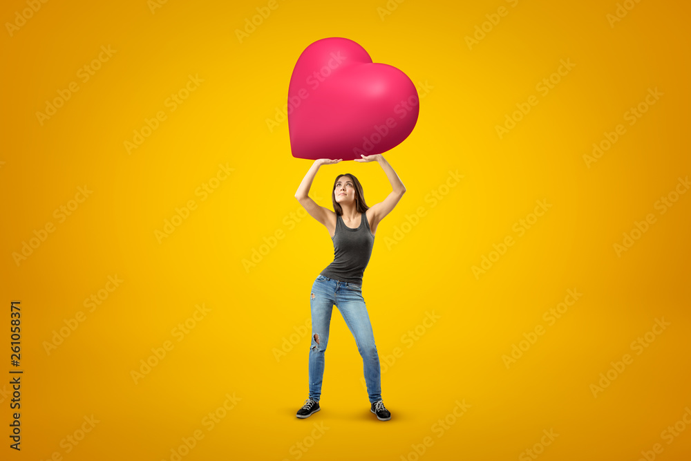 Young brunette girl wearing casual jeans and t-shirt holding big pink heart above on yellow background Young brunette girl wearing casual jeans and t-shirt holding big pink heart above on yellow background