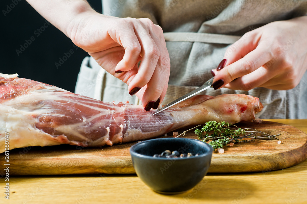 The chef cuts a raw leg of lamb on a wooden chopping Board. Rosemary, thyme, black pepper. Black background, side view, space for text