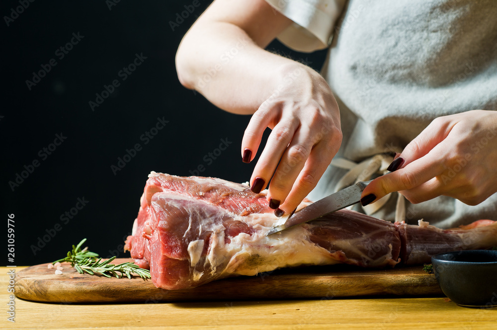 The chef cuts a raw goat leg on a wooden chopping Board. Rosemary, thyme, black pepper. Black background, side view, space for text