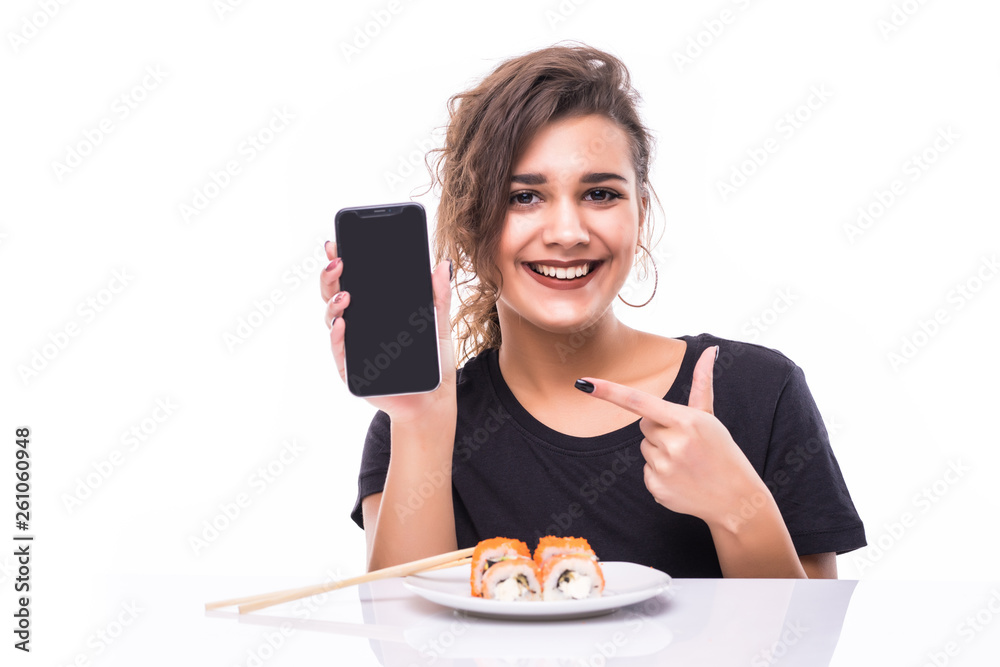 Smiling young woman showing blank screen mobile phone eating sushi at the table isolated over white background Smiling young woman showing blank screen mobile phone eating sushi at the table isolated over white background