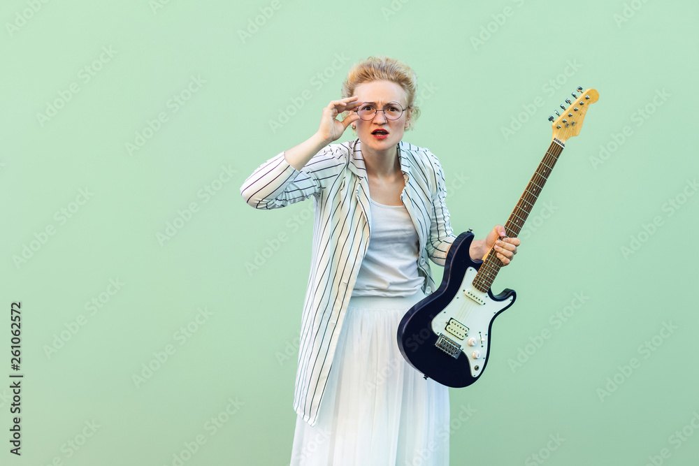 Portrait of young blonde woman in white shirt and striped blouse, electric guitar standing, holding eyeglasses and looking attentive at camera. indoor studio shot isolated on light green background.