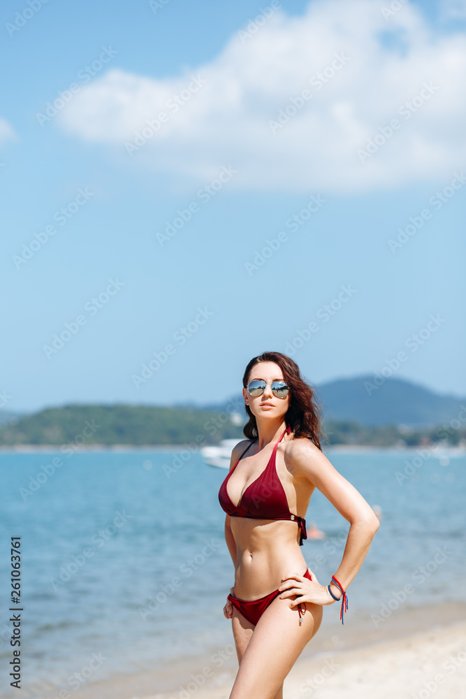 Beautiful brunette girl in a red bikini swimsuit and sunglasses posing on the beach on a Sunny day. Portrait of a woman on vacation against the sea.
