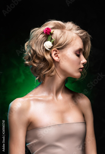 Beautiful young blonde girl with naked shoulders, flowers braided into her hair and oily body poses on a dark green gradient background. Clean, healthy skin. Nude make-up. Close up