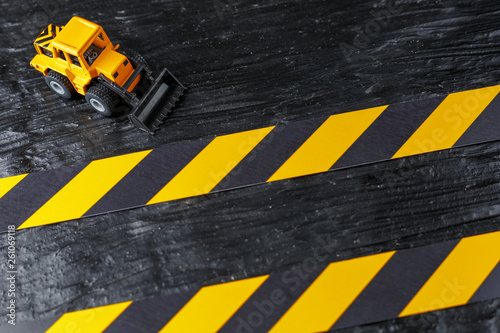 Yellow toy bulldozer on a black background texture. Black and yellow fence tape against the background of the CONCEPT