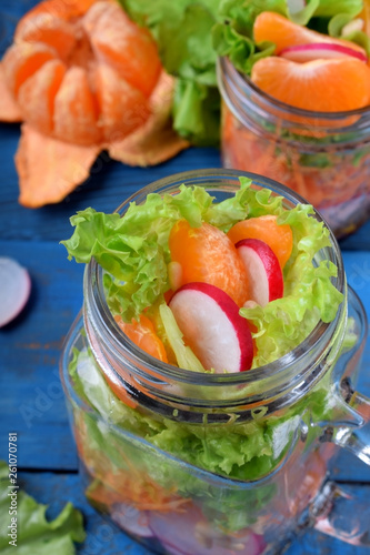 Layered salad in a jar with mandarins, radish, lettuce leaves, carrot and dressing against the blue background