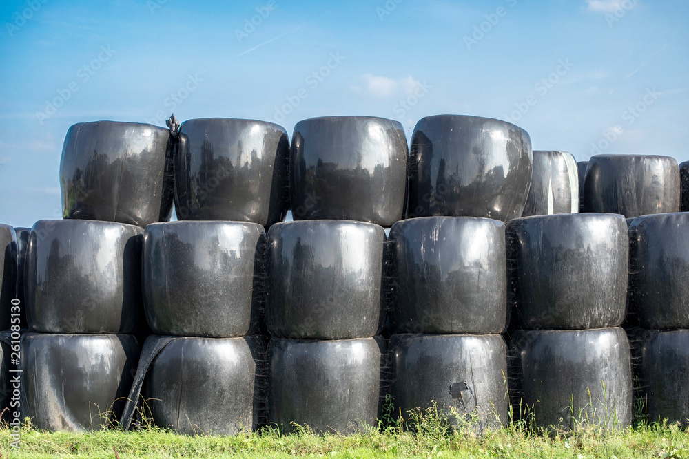Stack of bales of hay, piled up, wrapped in black plastic, on green grass under blue sky.